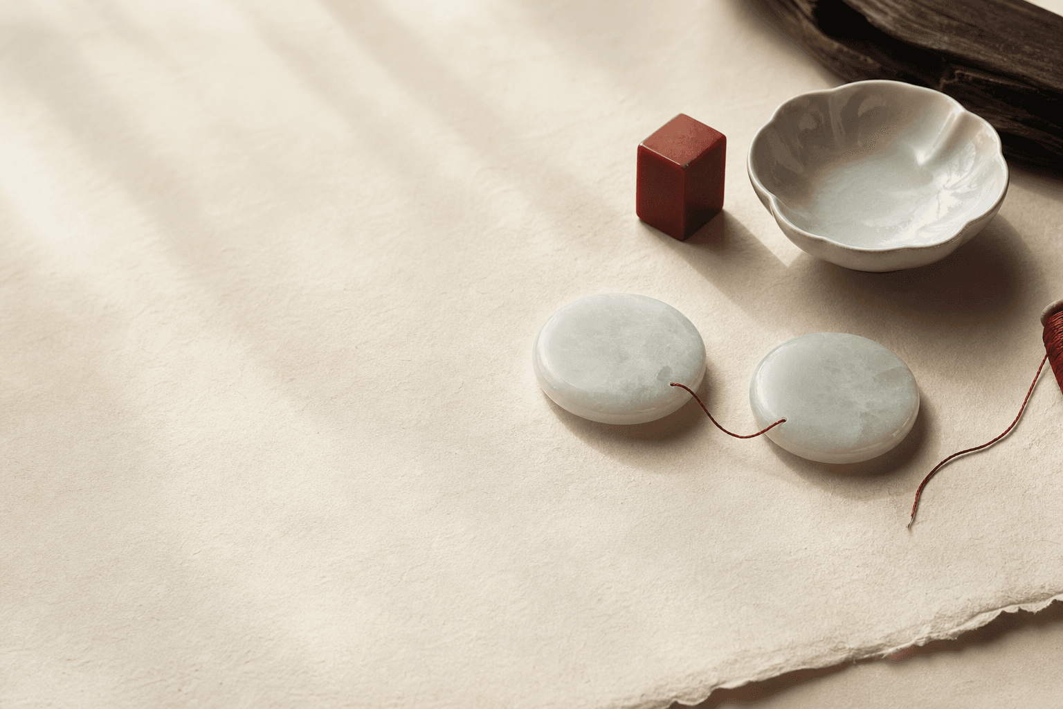 Two pale jade circles joined by red silk thread on handmade paper beside a porcelain dish